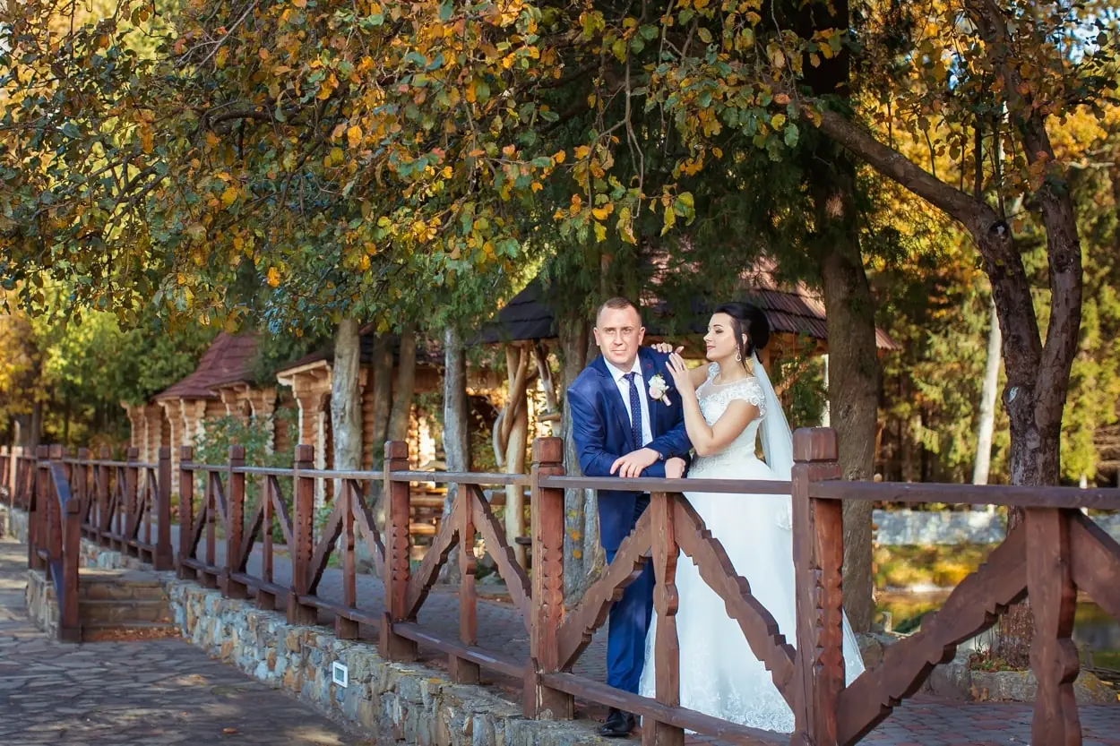 Молодята позують на фоні осінніх дерев біля дерев'яної огорожі на березі озера, професійна зйомка від RoStudio у Львові.