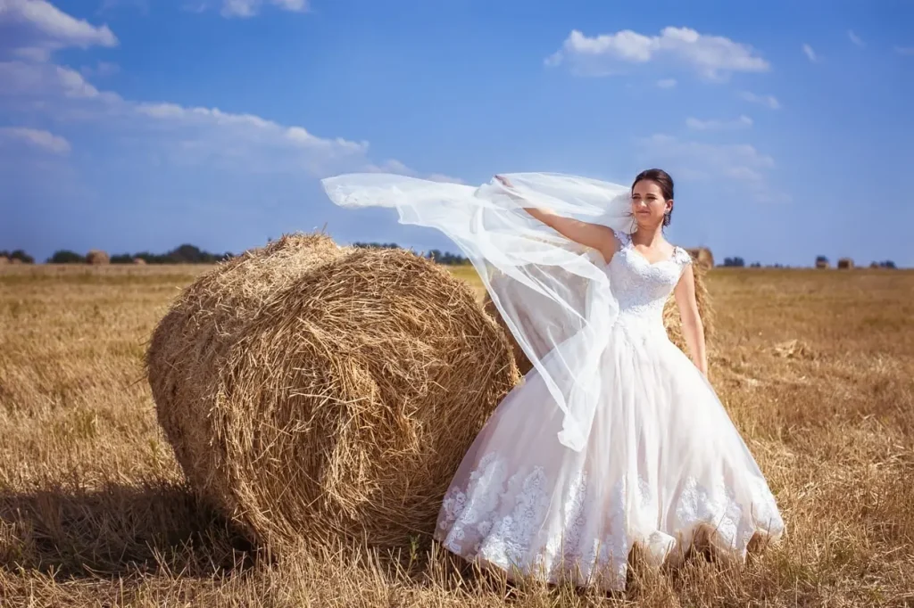 Наречена у білій сукні з фатою, що розвівається на вітрі, весільна фотосесія на полі серед сіна біля Львова від RoStudio.